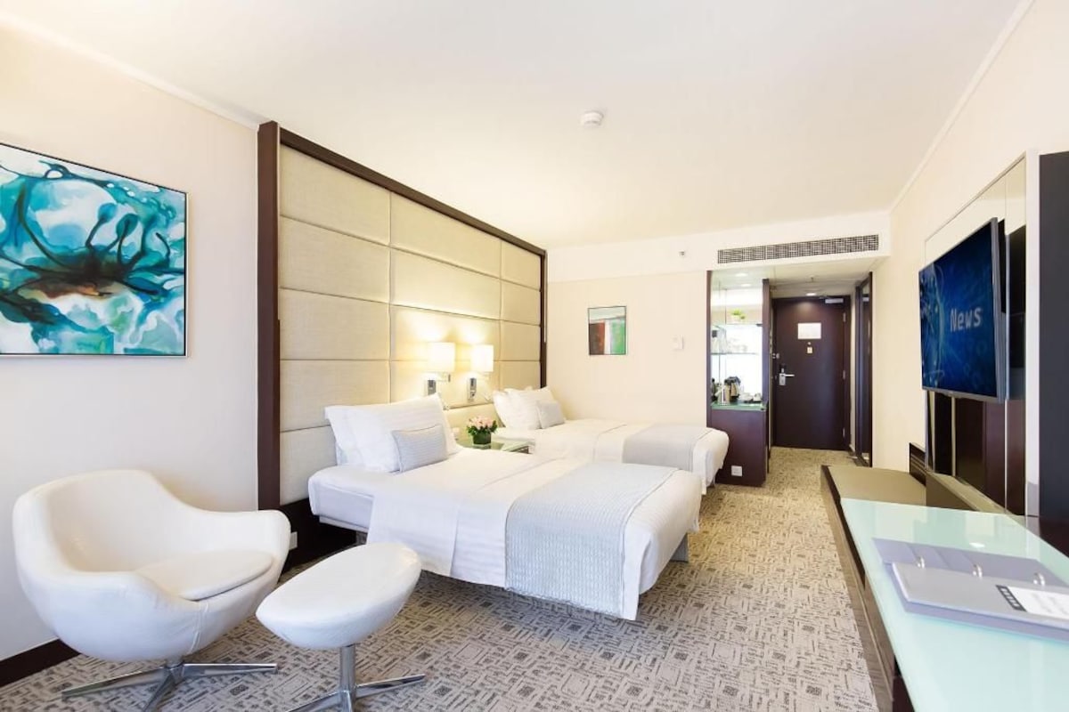 A hotel room with a bed, two white chairs, a desk, a TV, and artwork on the wall.