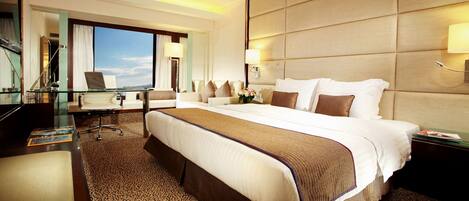 A hotel room with a large bed, a desk with a laptop, a chair, and a view of the sky.