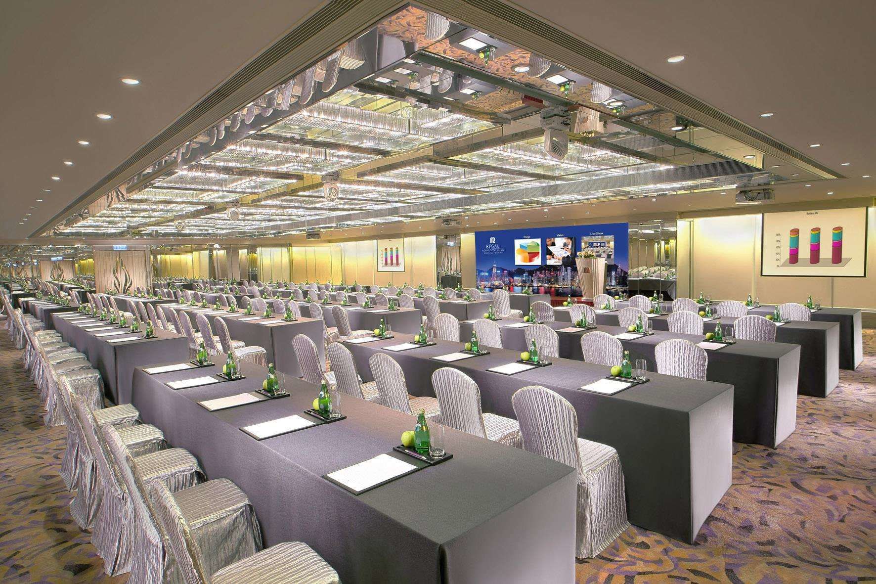 A conference room with rows of tables, chairs, and a large screen at the front.