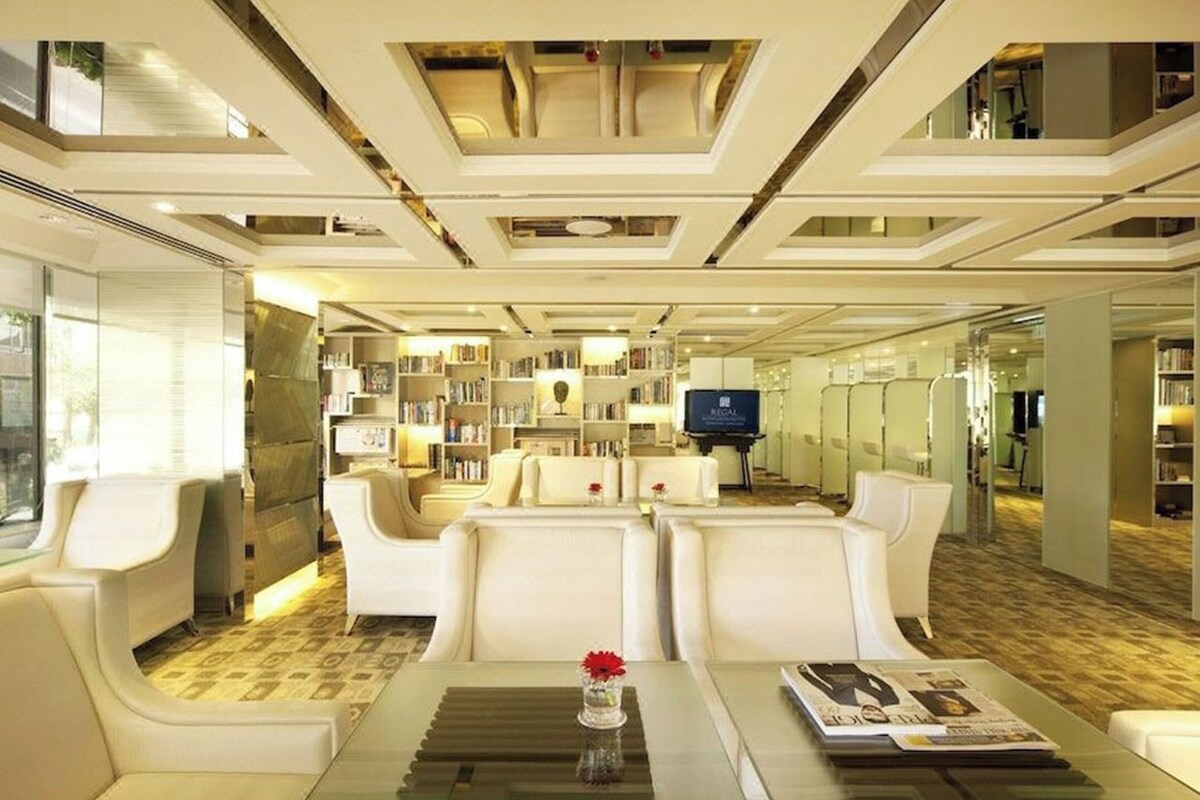 A modern hotel lobby with a central seating area, a large bookshelf, and a television.