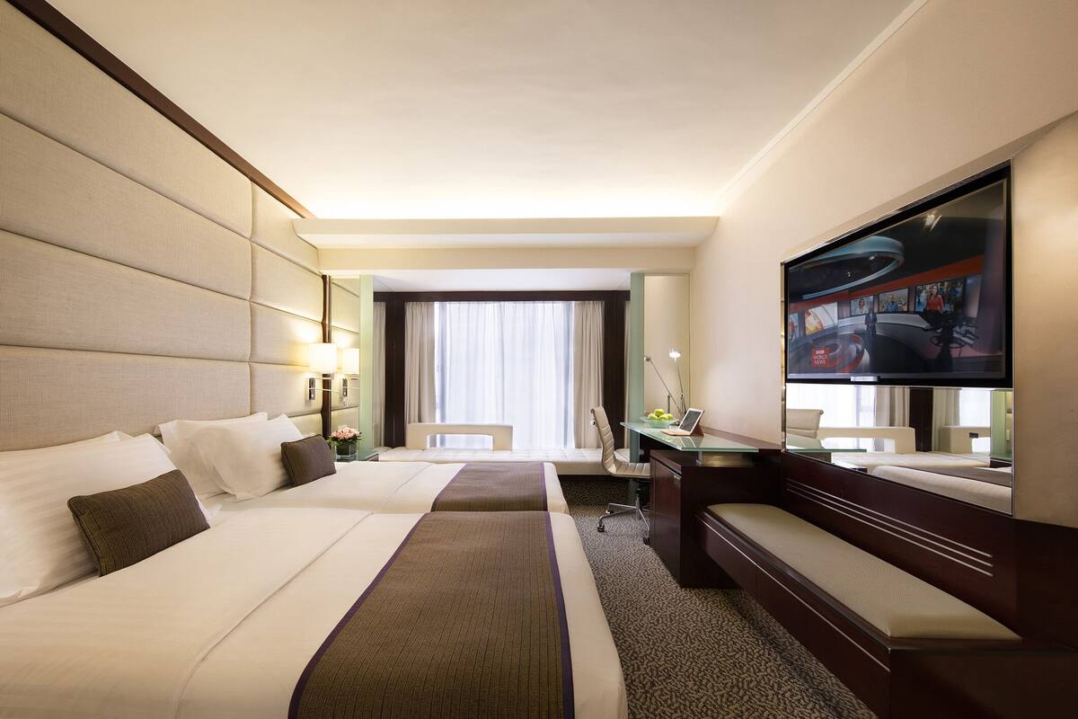 A hotel room with two beds, a desk, a TV, and a large window with curtains.