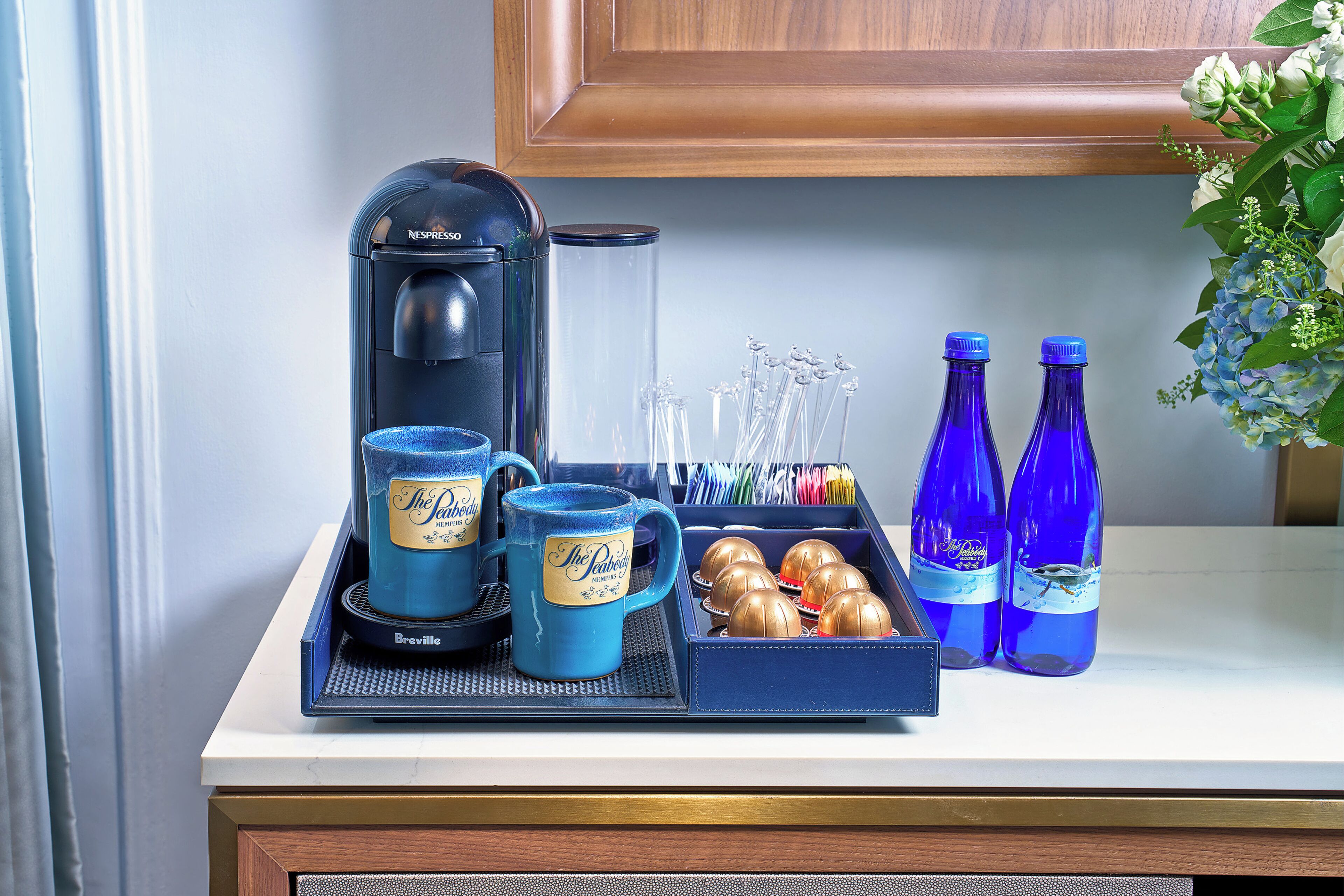 A coffee station with a Nespresso machine, two blue mugs, a tray with a coffee pot, and two bottles of water.
