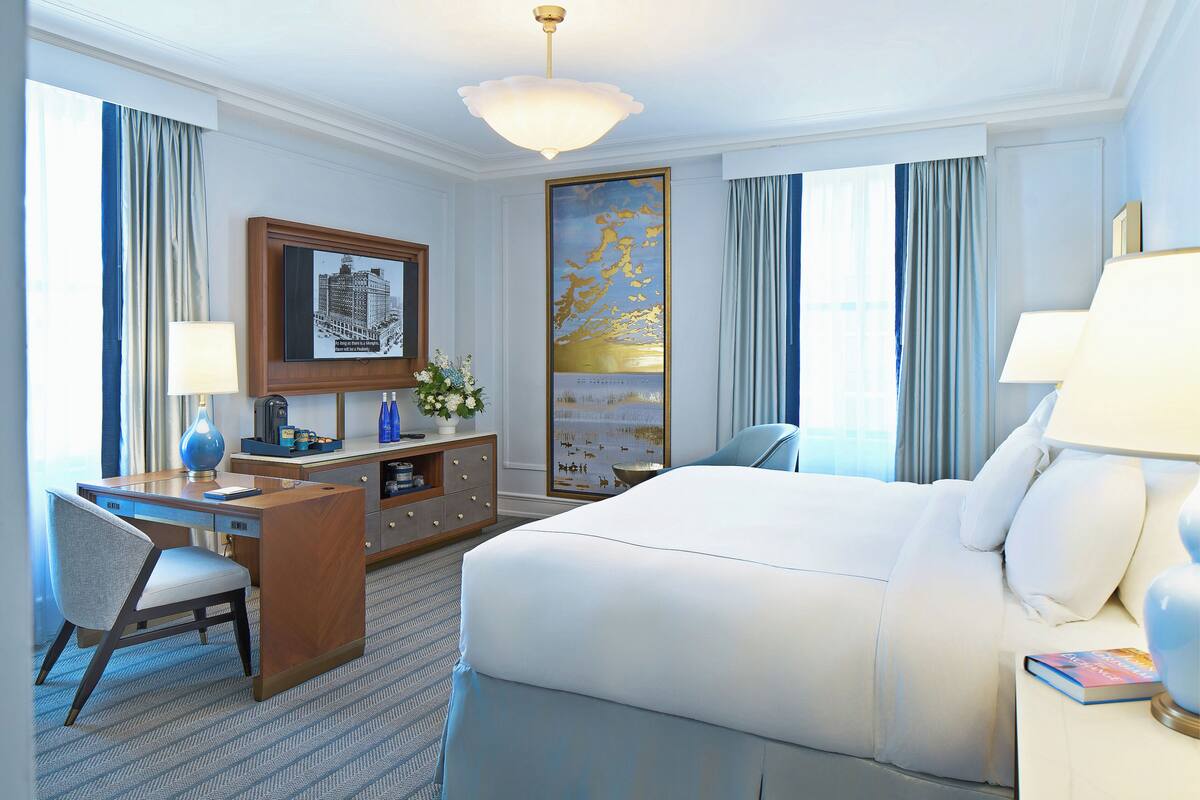 Ein Hotelzimmer mit einem großen Bett, einem Schreibtisch und einem Sessel. An der Wand hängt ein Bild und ein Fenster mit Vorhängen.