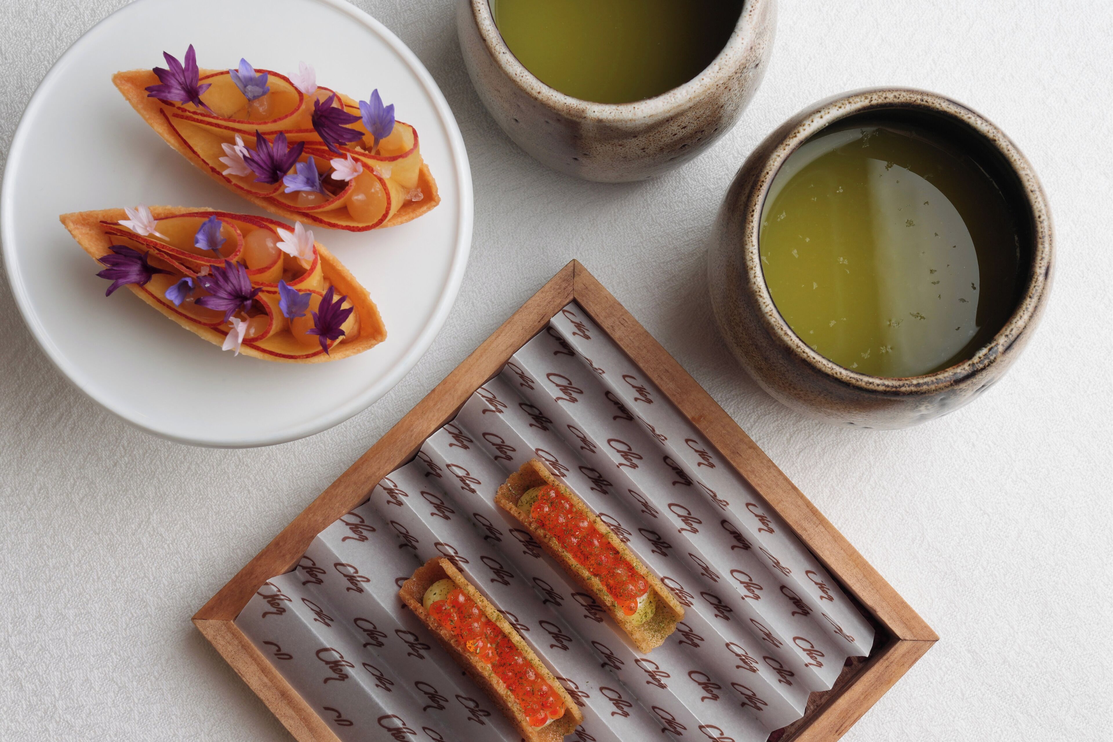 A plate with two pastries, a wooden box with a patterned surface, and two ceramic cups with green liquid.