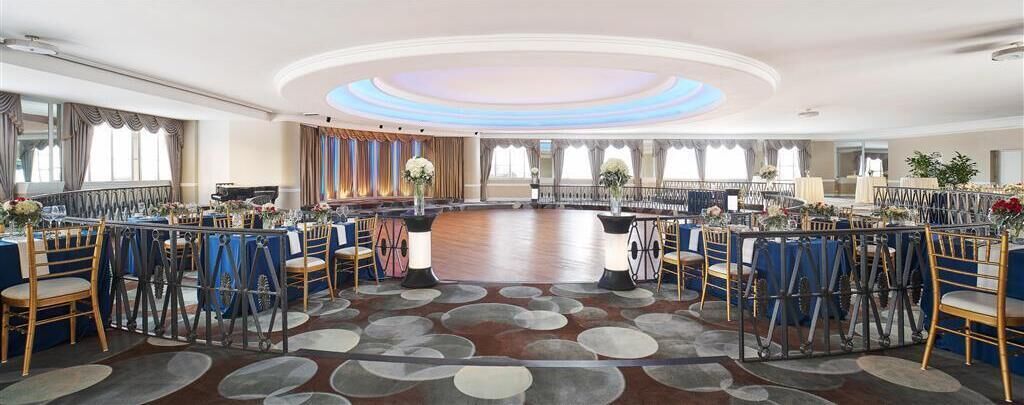 Uma grande sala de banquetes circular com chão de madeira e cadeiras azuis e brancas dispostas em volta de mesas.