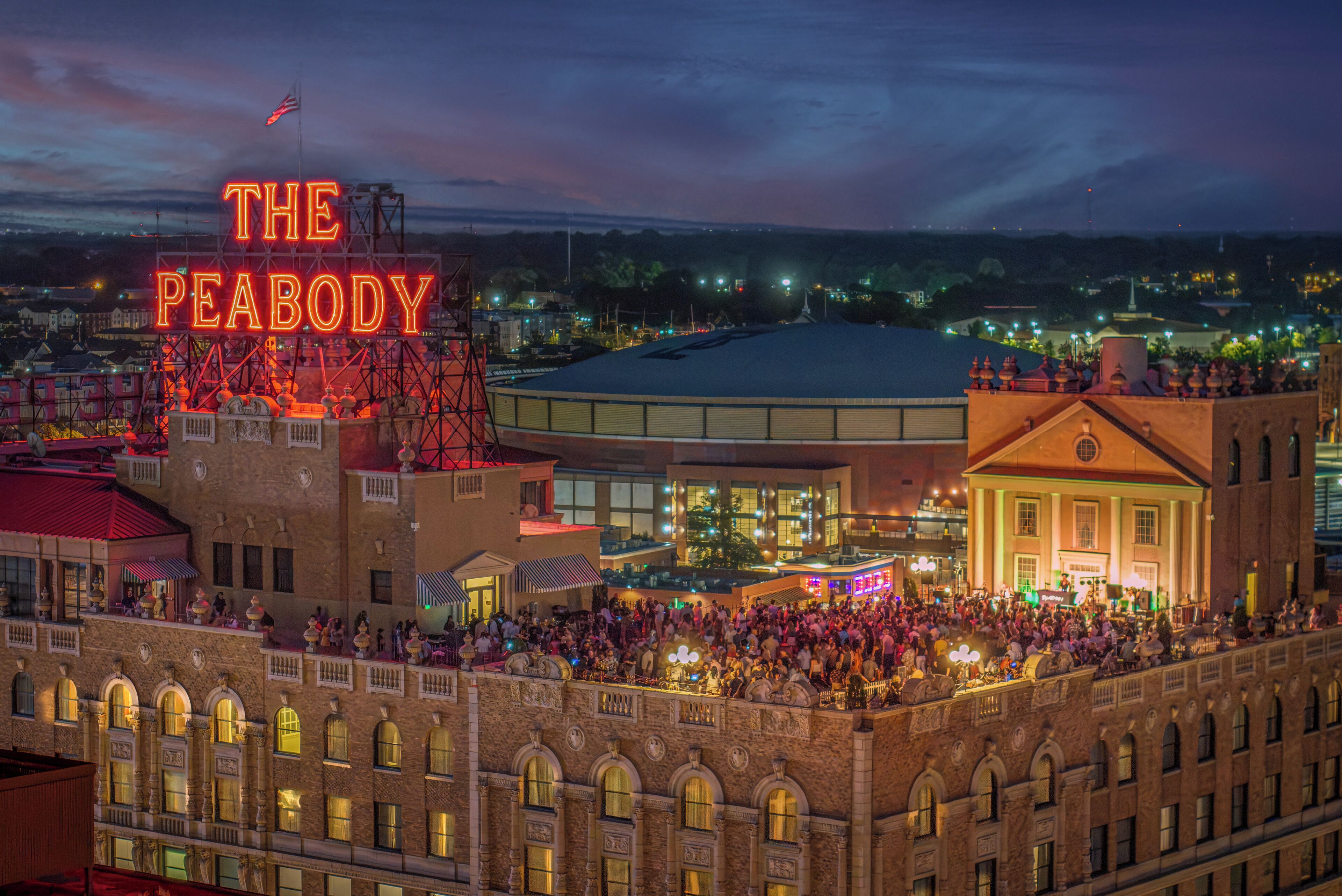 A historic building with a large crowd gathered on its upper level, a prominent neon sign reading "THE PEABODY," and a flag on top.