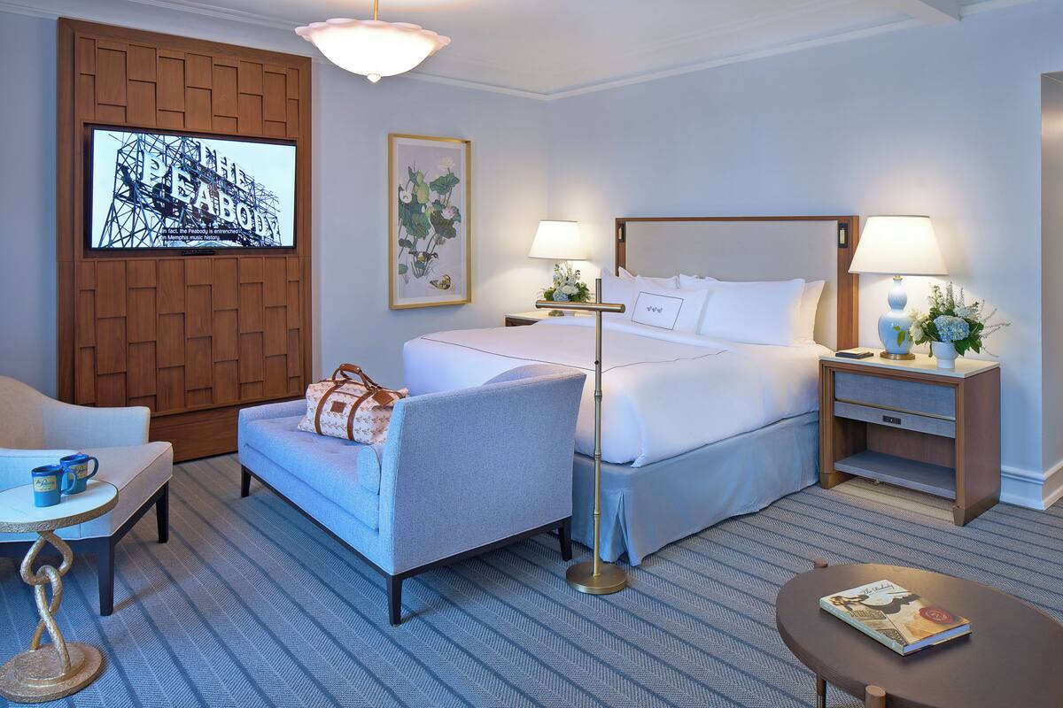 Ein Hotelzimmer mit einem Bett, zwei Nachttischen, einem Sessel, einem kleinen Tisch und einem an der Wand montierten Fernseher.