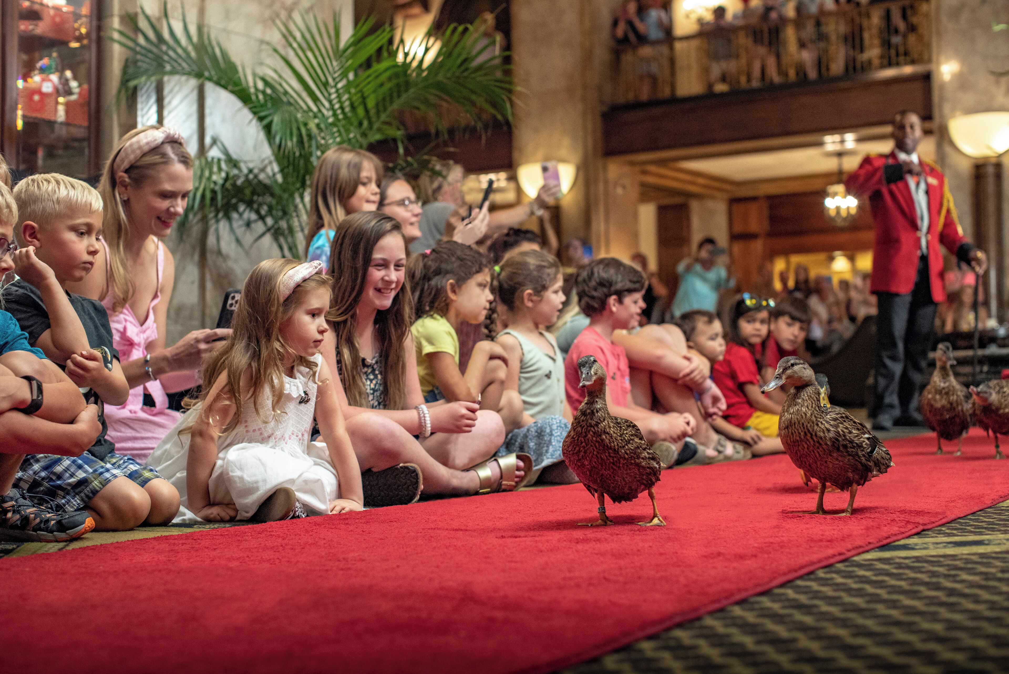 A group of children sitting on a red carpet in a lobby with a red carpet, a plant, and a person in a red uniform.
