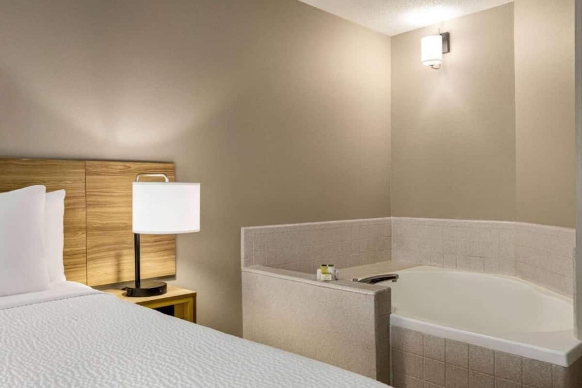 A hotel room with a bed, a nightstand with a lamp, a bathroom with a bathtub, and a wall-mounted light fixture.