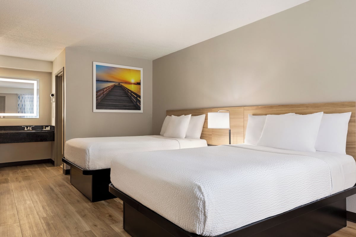A hotel room with two beds, a wooden headboard, a painting of a sunset, a bathroom with a mirror and sink, and a wooden floor.