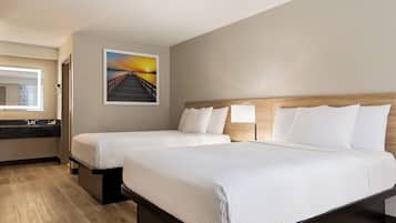 A hotel room with two beds, a wooden headboard, a painting of a sunset, a bathroom with a mirror and sink, and a wooden floor.