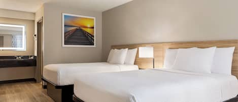 Habitación de hotel con dos camas, cabecero de madera, un cuadro de una puesta de sol, baño con espejo y lavabo, y suelo de madera.