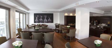 Une salle à manger d’hôtel avec de nombreuses tables et chaises, une grande statue de Bouddha accrochée au mur et une vue sur la ville grâce à de grandes fenêtres.