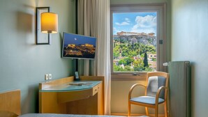Habitación de hotel con cama, escritorio con ordenador, silla, televisor mostrando una vista urbana y ventana con vista a un edificio histórico.