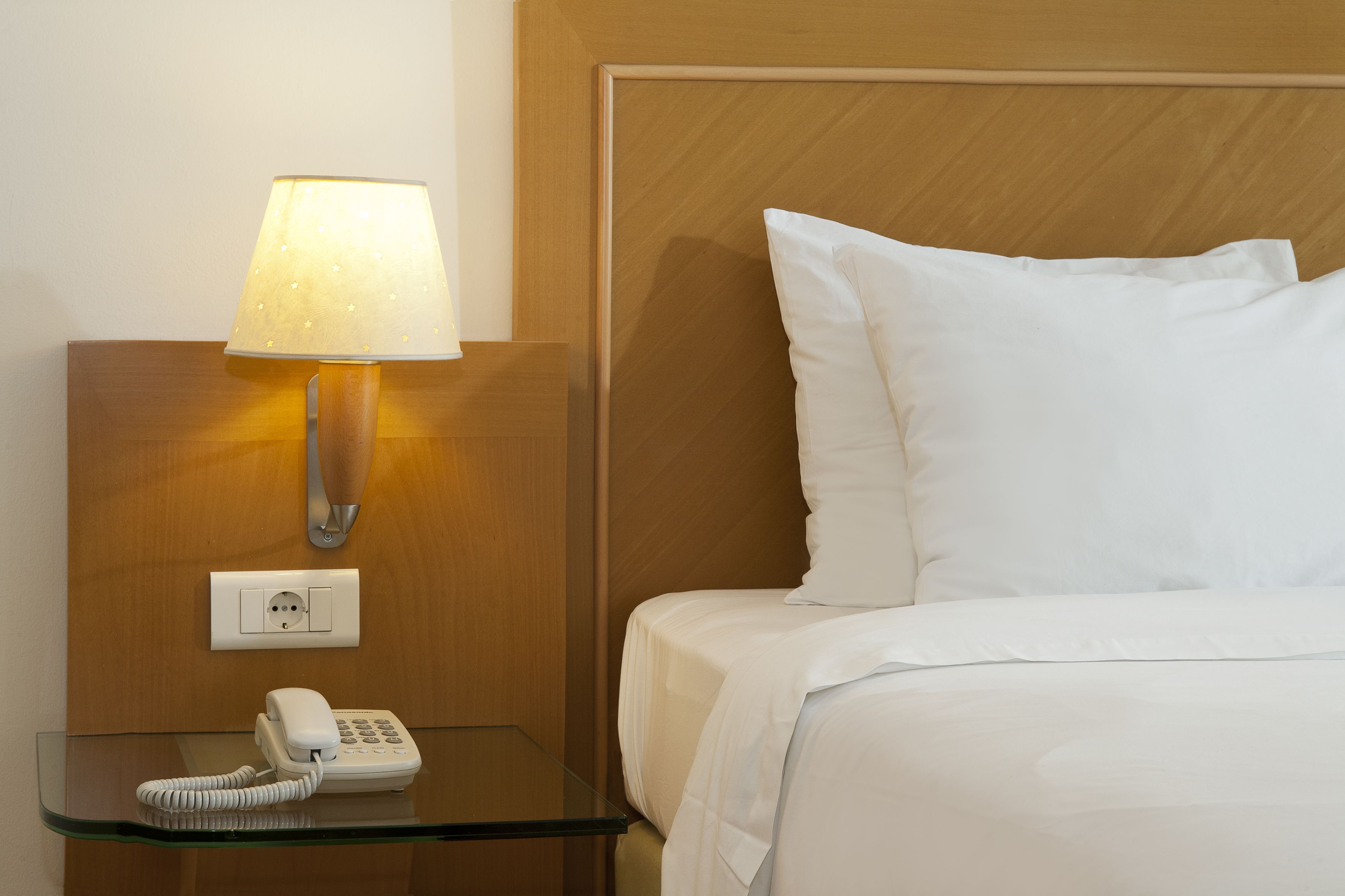 A hotel room with a bedside table, a lamp, a phone, and a bed with white bedding.