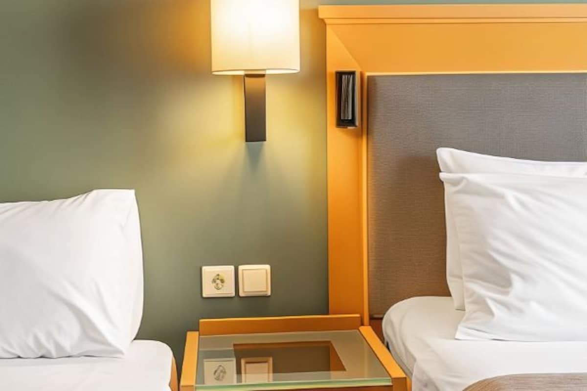 Habitación de hotel con dos camas, una mesita de noche, una lámpara y un interruptor de luz en la pared.