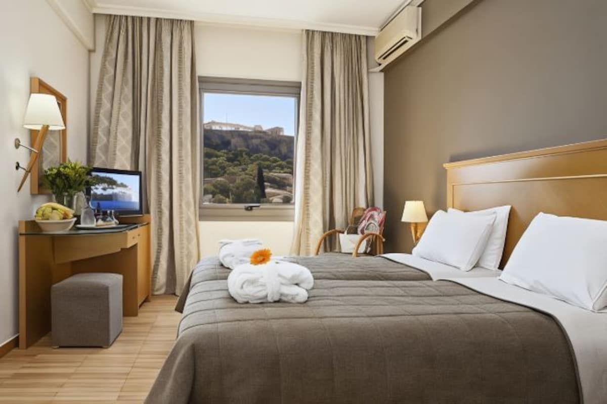 Habitación de hotel con una cama grande, un escritorio con computadora, una ventana con vista a un paisaje y una pequeña mesa con una planta.