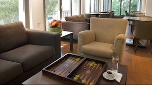 Habitación de hotel moderna con un ventanal grande, un sofá, una silla, una mesa de centro con un juego de mesa y una zona de comedor al fondo.