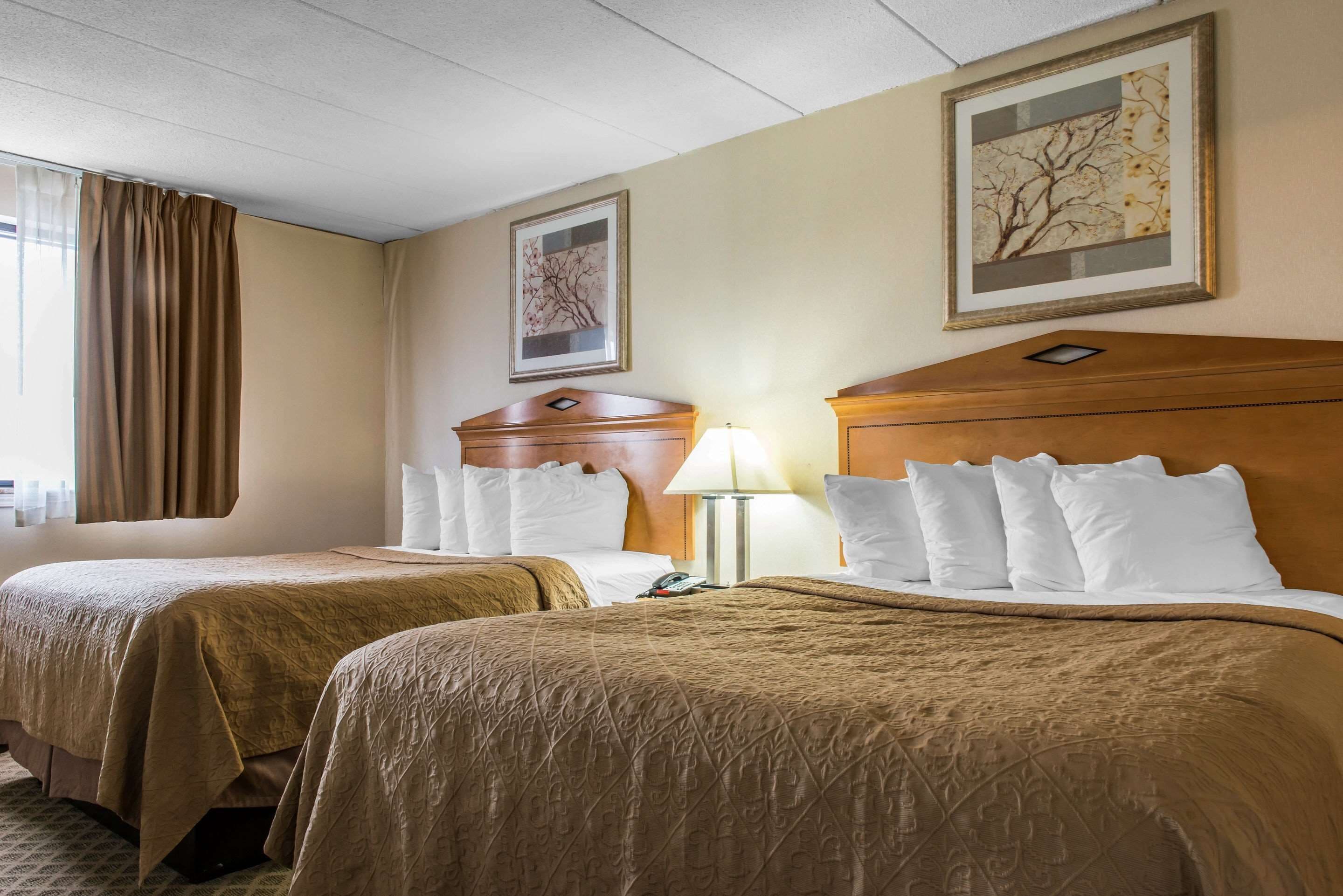Une chambre d’hôtel avec deux lits, des murs de couleur beige, une tête de lit en bois, une table de chevet, une lampe et des œuvres d’art encadrées accrochées aux murs.