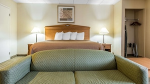A hotel room with a bed, two bedside lamps, a wooden headboard, a framed picture on the wall, and a coat rack with hangers.