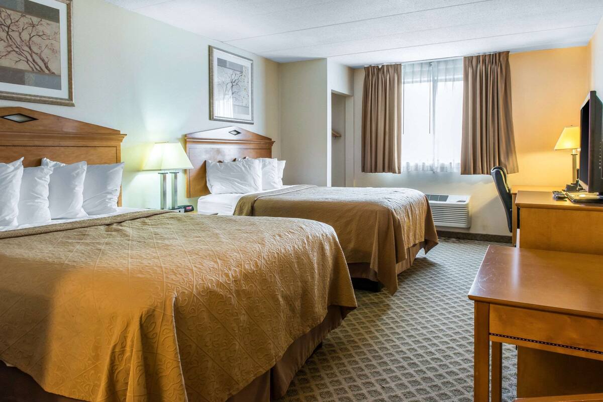 Quarto de hotel com duas camas, uma escrivaninha, uma cadeira, uma televisão e uma janela com cortinas.