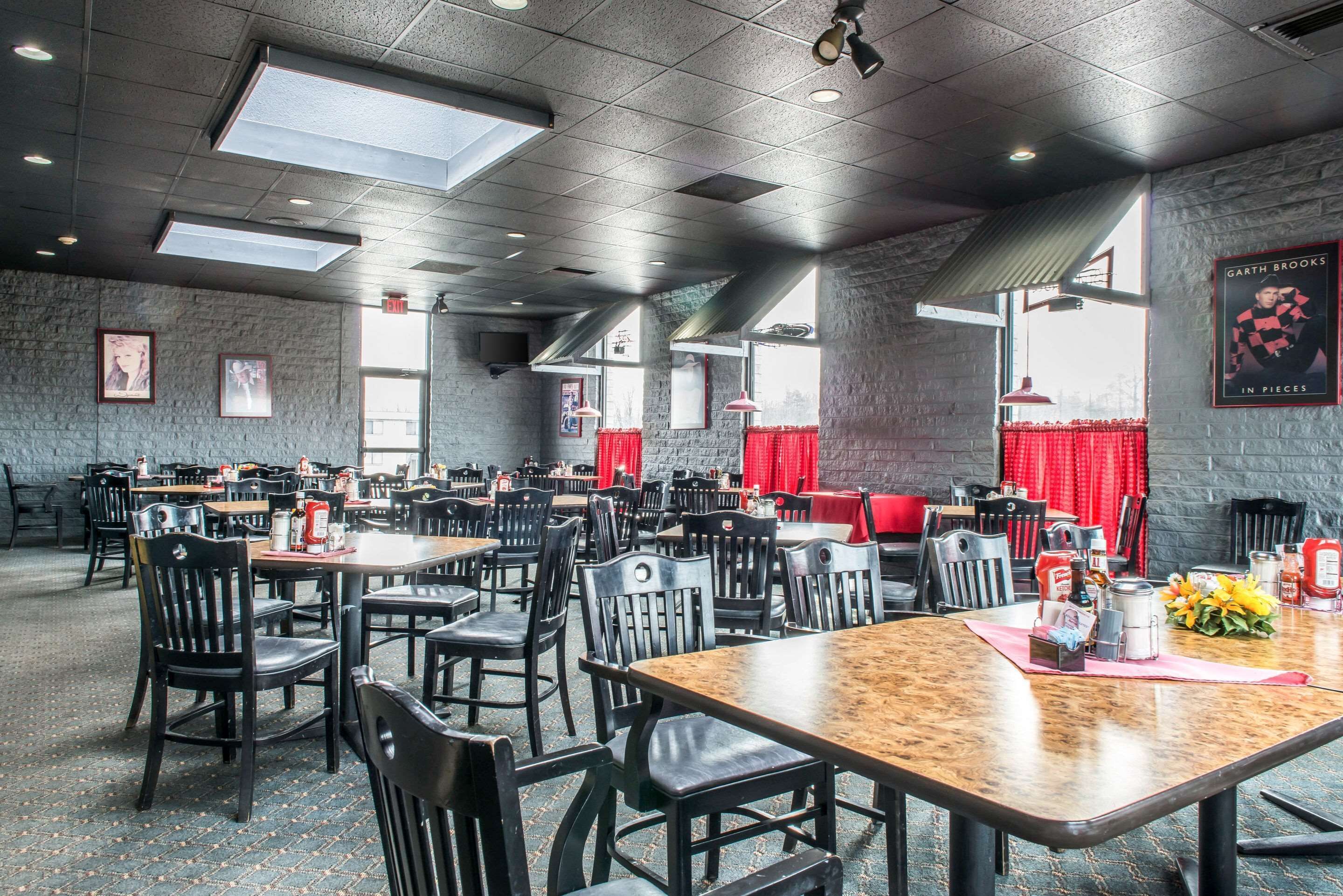 A spacious restaurant with multiple tables and chairs, large windows with red curtains, and a brick wall.