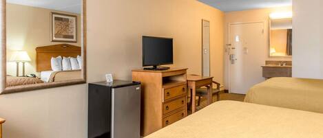 A hotel room with a bed, a desk with a television, a refrigerator, a dresser, and a mirror.