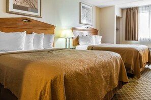 A hotel room with two beds, each with a wooden headboard, white bedding, and a patterned bedspread. There is a nightstand with a lamp, a framed picture on the wall, and a window with curtains.