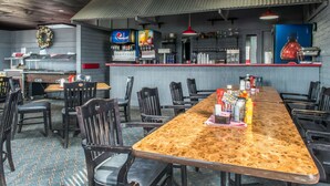 A restaurant with a long wooden table, black chairs, a bar area with a Pepsi machine, and a red hanging light.