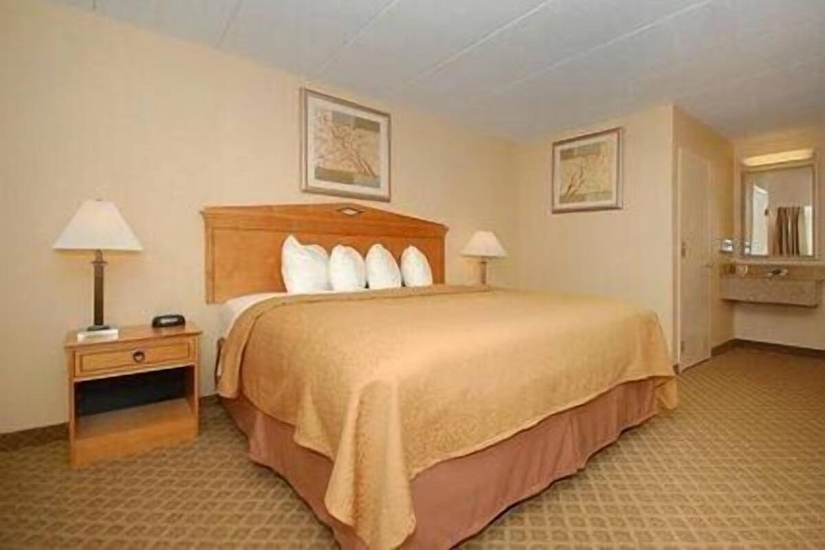 Quarto de hotel com uma cama, duas mesas de cabeceira, um criado-mudo, um espelho e duas fotografias emolduradas na parede.