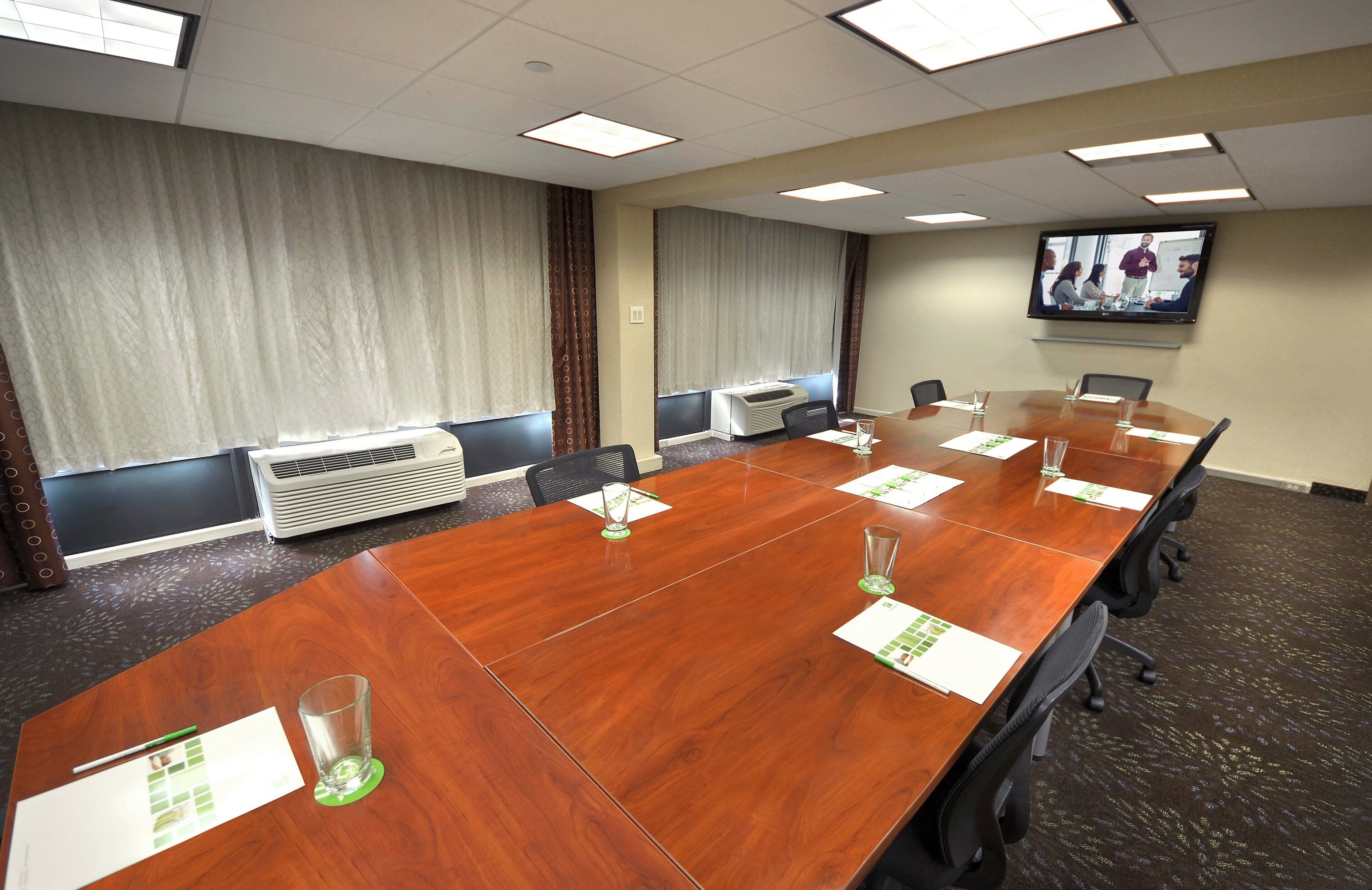 A conference room with a long wooden table, chairs, a large flat-screen TV on the wall, and an air conditioning unit.