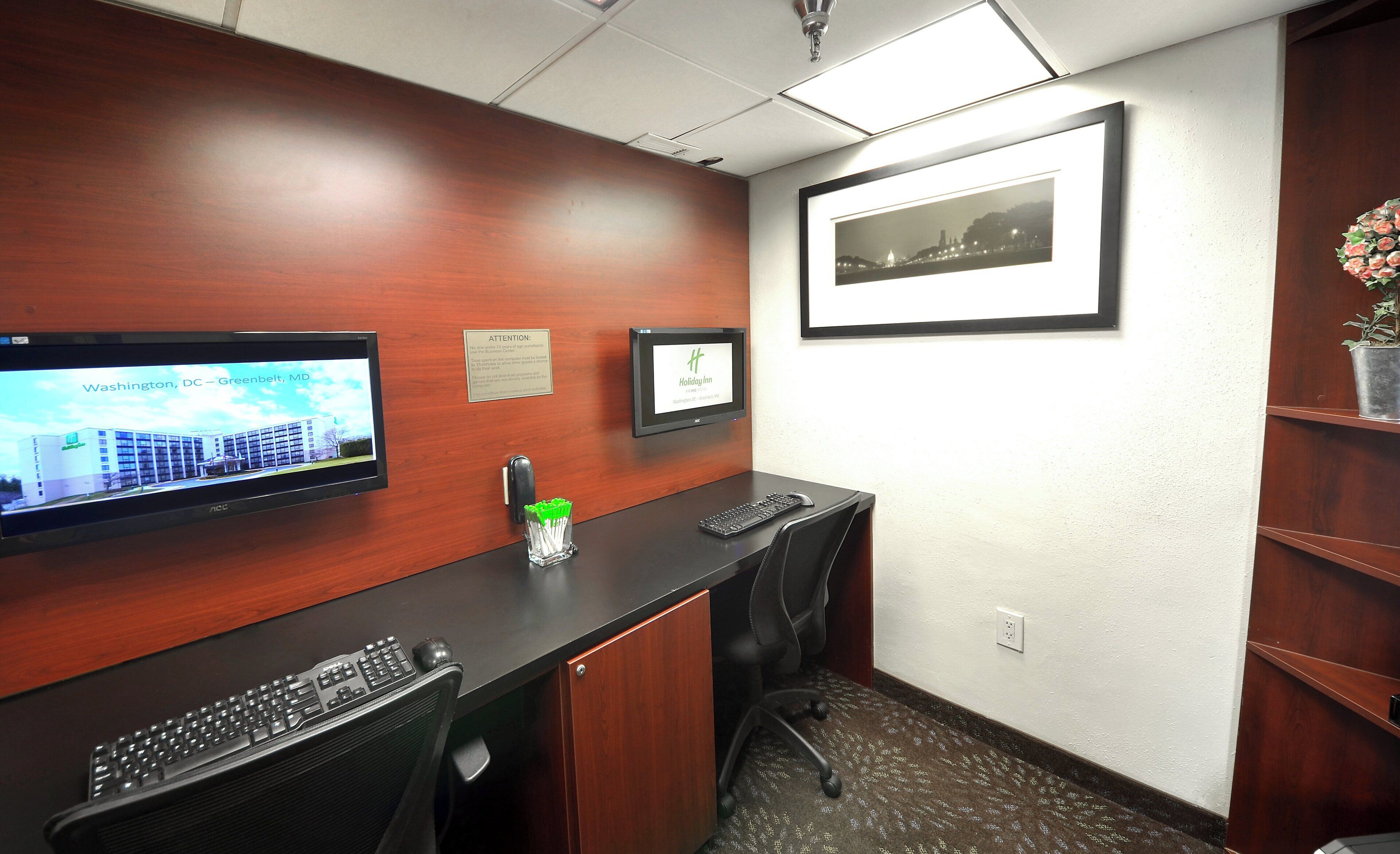 A hotel reception desk with a monitor displaying a cityscape, a framed picture of a cityscape, and a small potted plant.