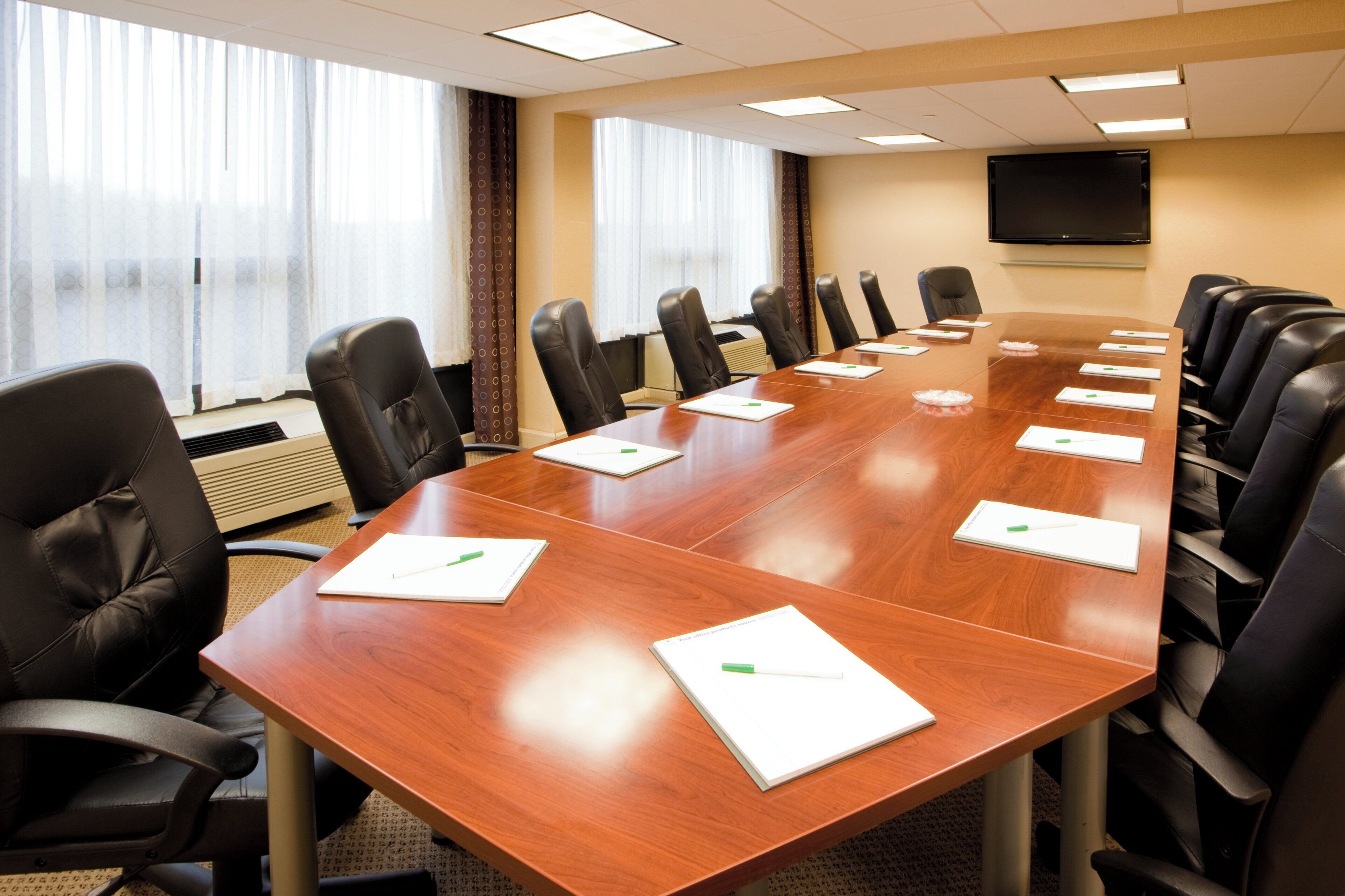 A conference room with a long wooden table, black leather chairs, and a flat-screen TV on the wall.