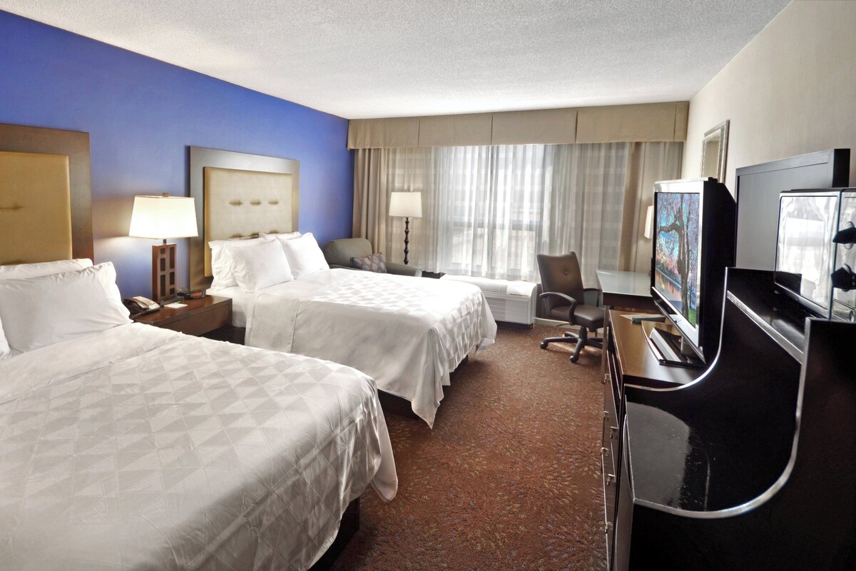 A hotel room with two beds, a desk with a TV, a chair, and a window with curtains.