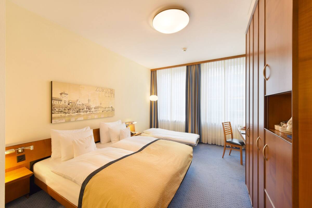 Quarto de hotel com duas camas, uma escrivaninha, uma cadeira, um guarda-roupa e uma janela grande com cortinas.