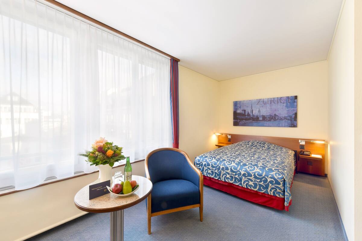 Quarto de hotel com cama, uma cadeira, uma mesa com um vaso de flores e um quadro na parede.