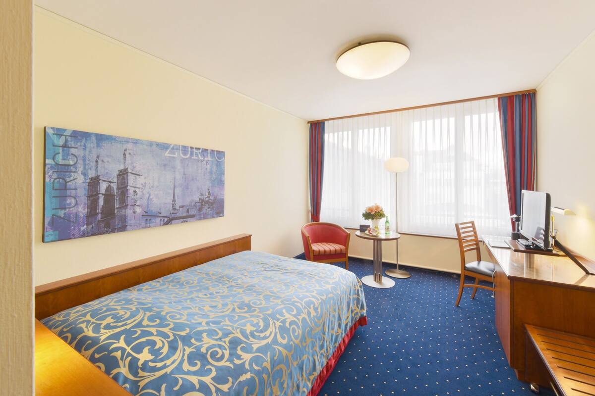 Quarto de hotel com cama, escrivaninha, cadeira, mesinha de centro com um vaso e um quadro na parede.