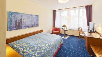 A hotel room with a bed, a desk, a chair, a small table with a vase, and a painting on the wall.