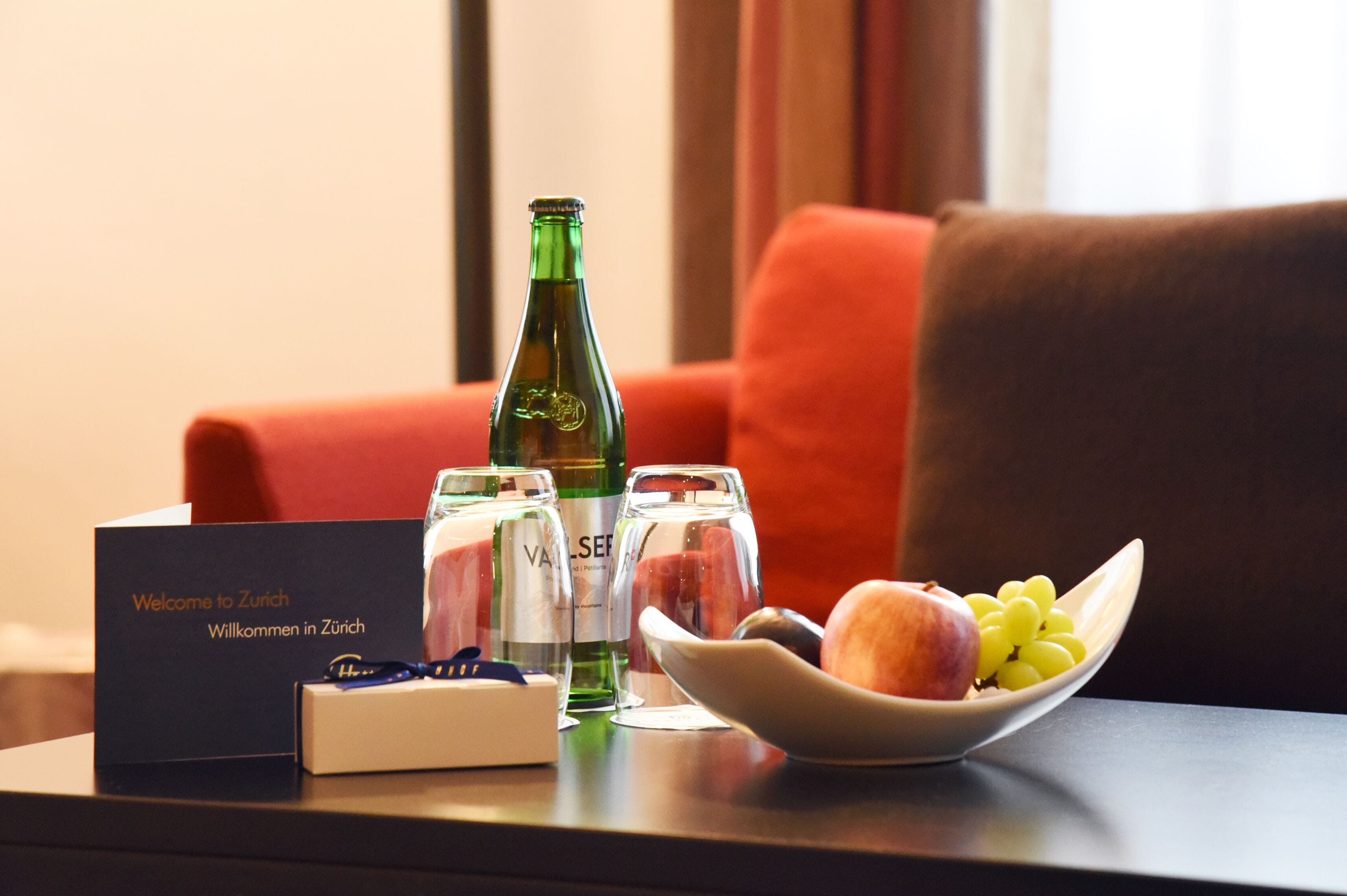 Un cartel de bienvenida, una botella, vasos y un cuenco con fruta sobre una mesa en una habitación de hotel.