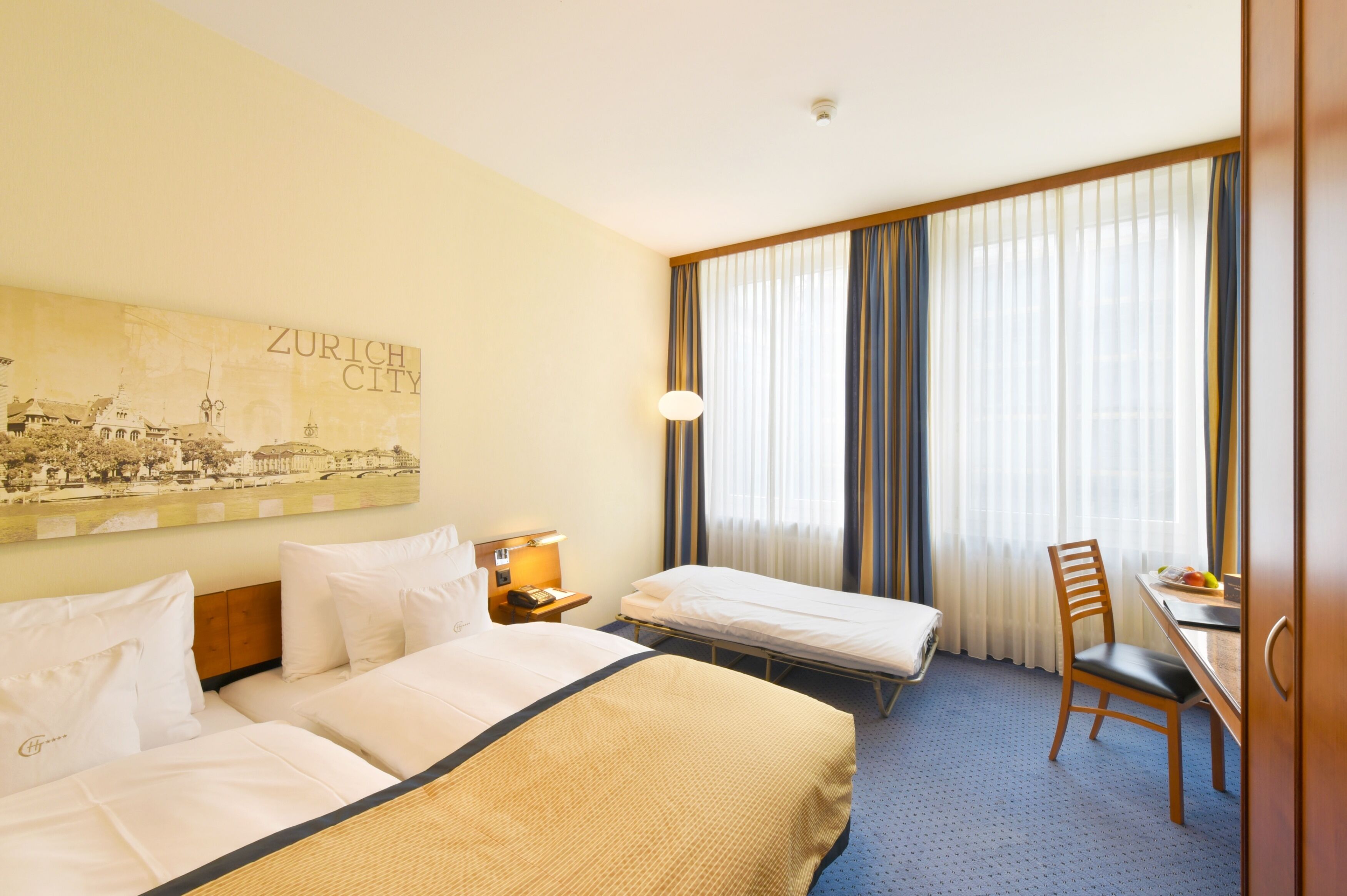 Habitación de hotel con dos camas, un escritorio, una silla, una lámpara y un ventanal grande con cortinas.