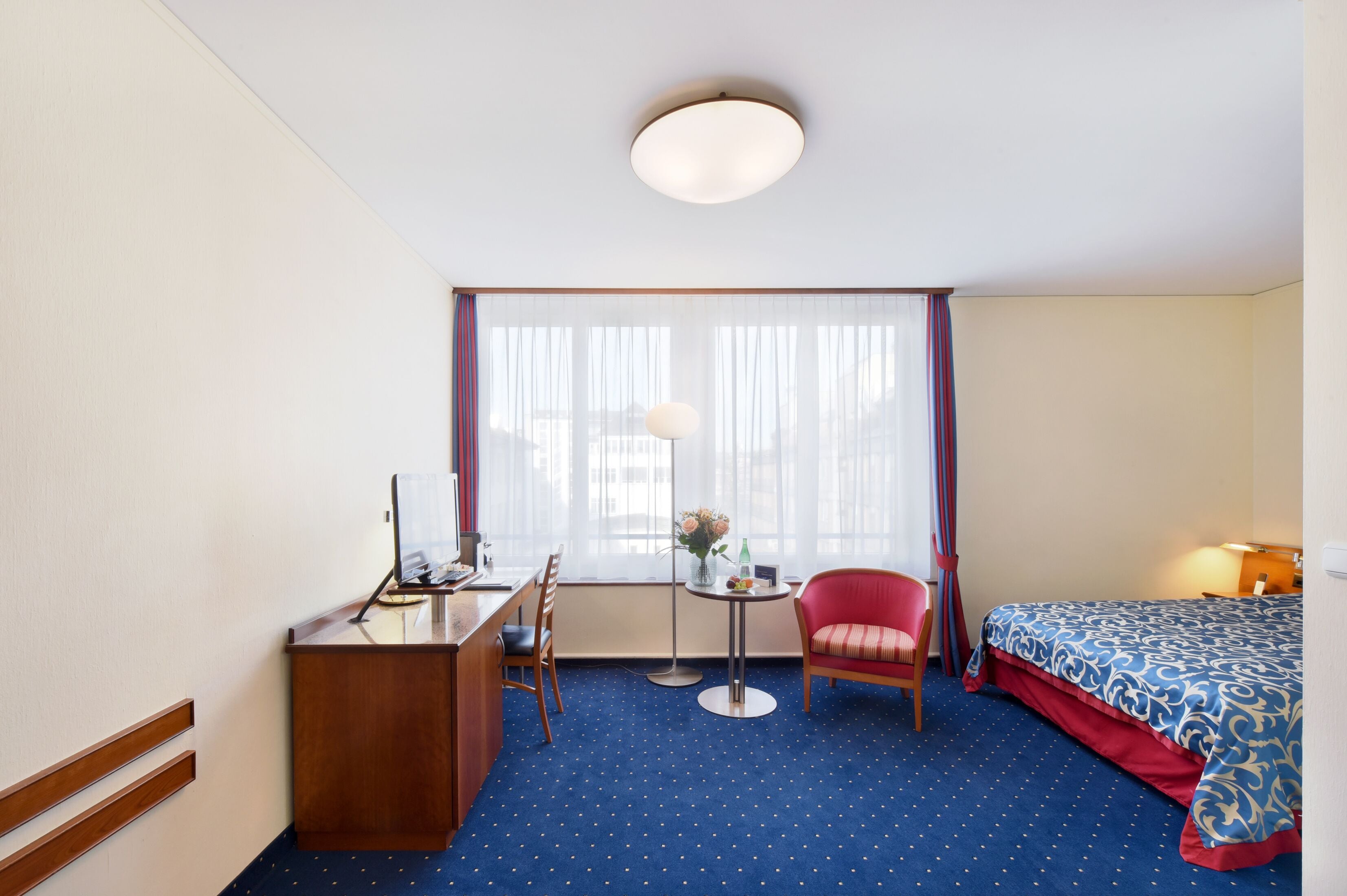 Ein Hotelzimmer mit Schreibtisch, Stuhl, einem kleinen runden Tisch und einem Bett mit gemustertem Überwurf.