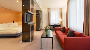 A hotel room with a bed, a red sofa, a coffee table with flowers, a TV, and a desk with a lamp. - Glärnischhof by Trinity (Zürich)