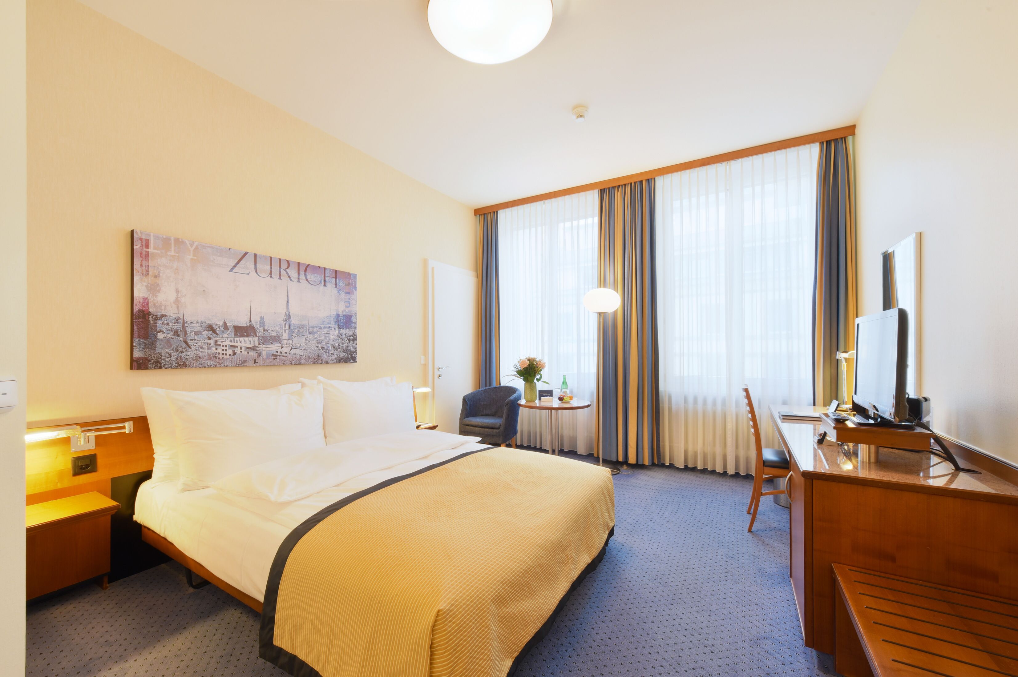 A hotel room with a large bed, a desk, a chair, a small table with a vase, and a painting of a cityscape.