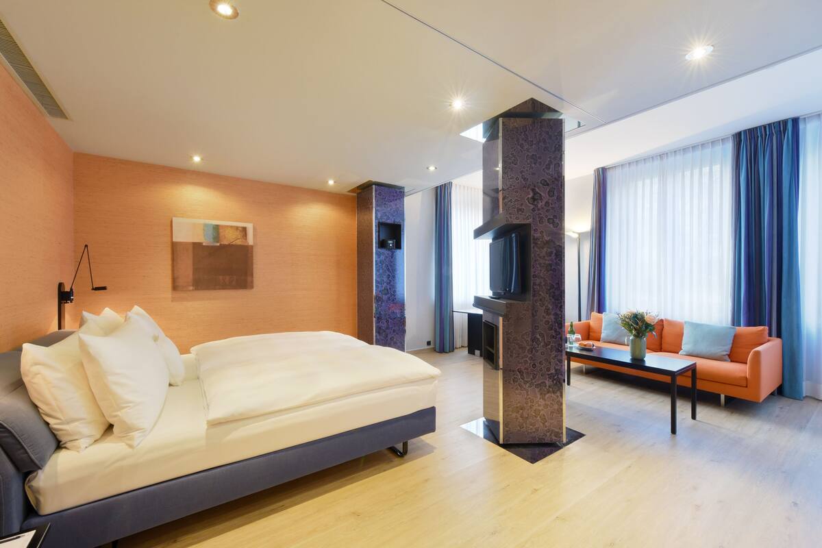Quarto de hotel moderno com uma cama grande, um sofá, uma mesa de centro e uma TV.