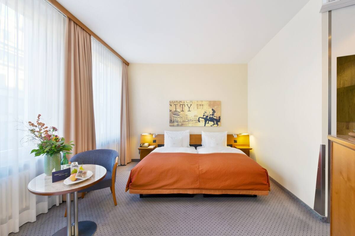 Quarto de hotel com cama, uma cadeira, uma pequena mesa com um vaso de flores e um quadro na parede.