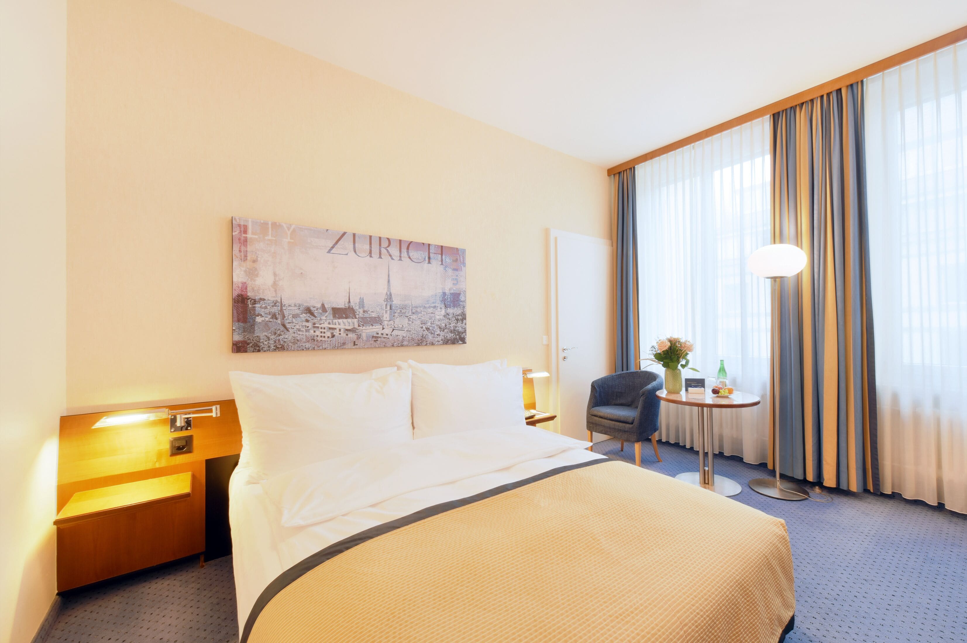 A hotel room with a large bed, a nightstand with a lamp, a chair, a small table with a vase and flowers, and a window with curtains.