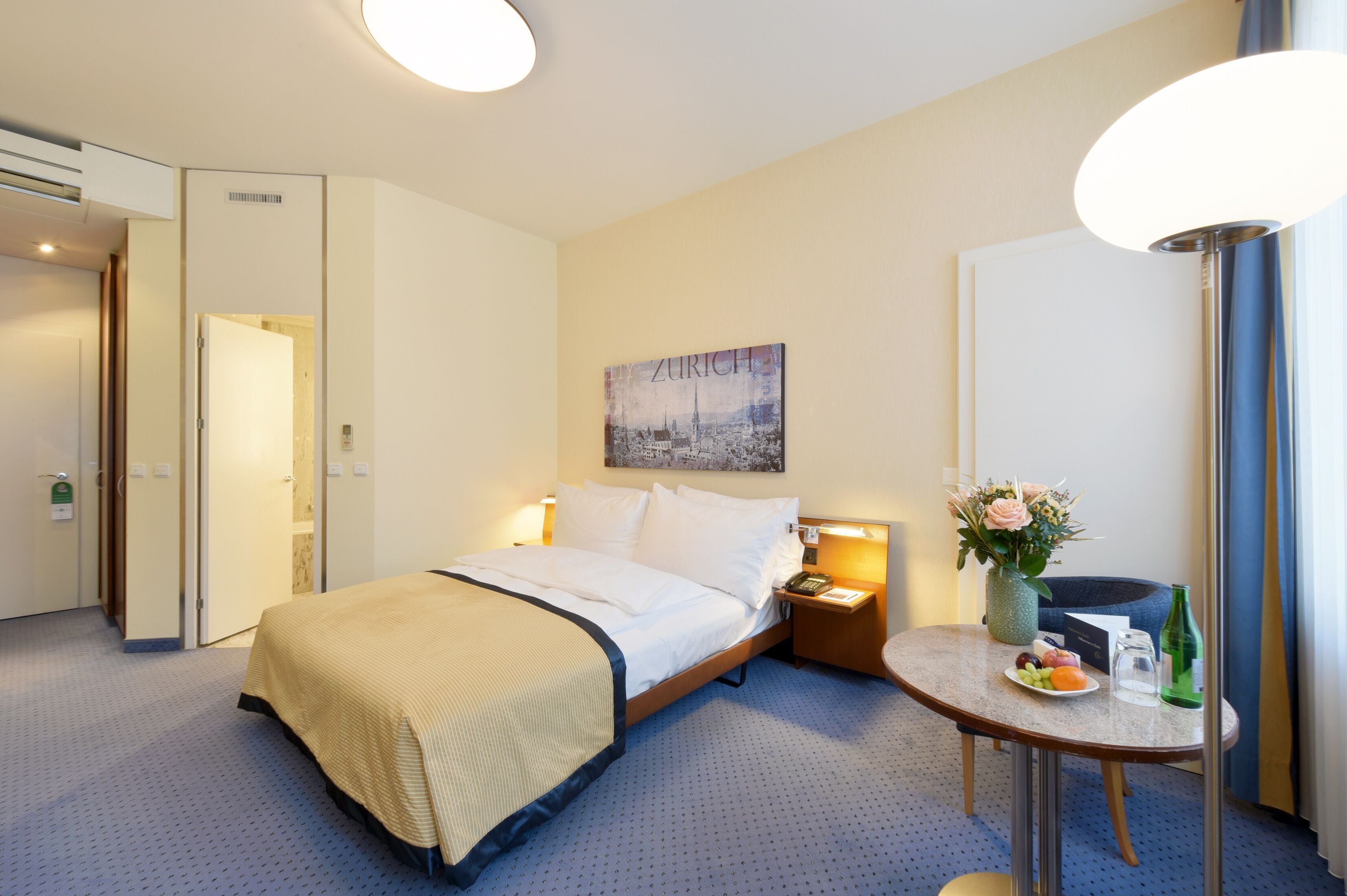 A hotel room with a bed, bedside table with a vase of flowers, a small table with fruit and a bottle, and a painting on the wall.