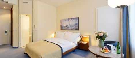 A hotel room with a bed, bedside table with a vase of flowers, a small table with fruit and a bottle, and a painting on the wall.