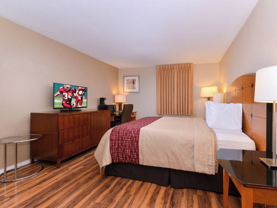 Quarto de hotel com cama, escrivaninha, televisão, cadeira, abajur e um quadro na parede.