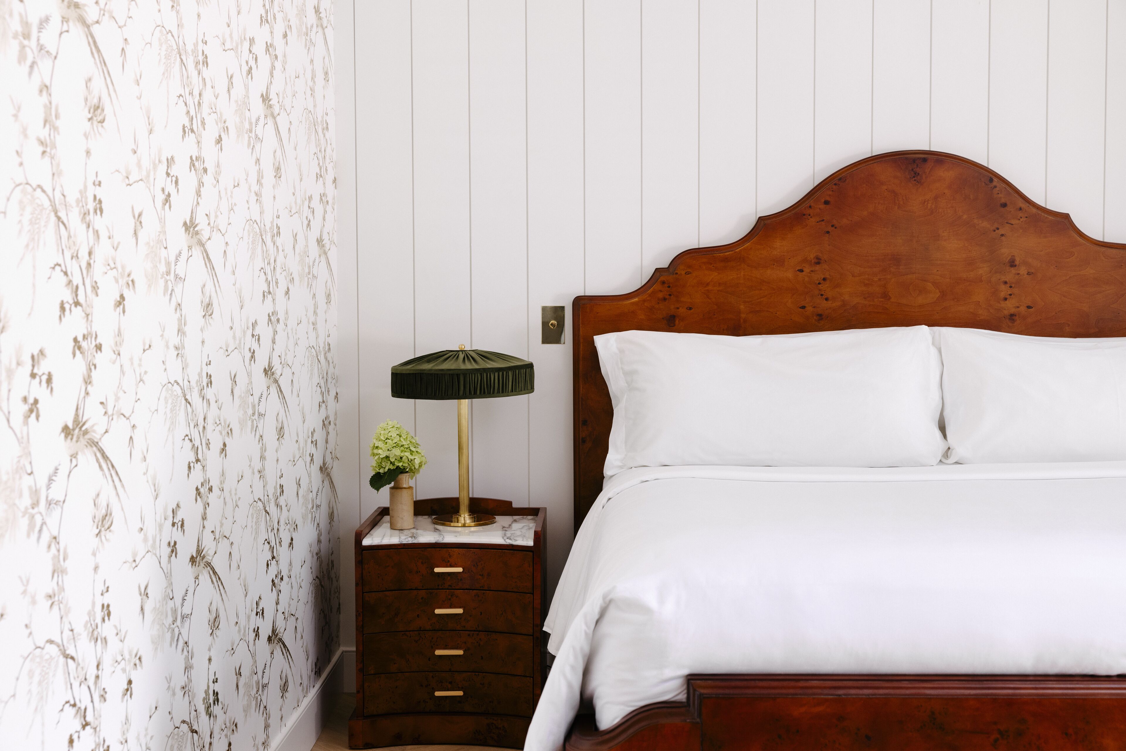 A neatly made bed with white linens, a wooden headboard, and a nightstand with a lamp and a small vase with flowers.